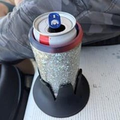 A black Steadi on a grandstand bench holding a canned drink in a glitter koozie, a spectator's leg beside it, at an outdoor race